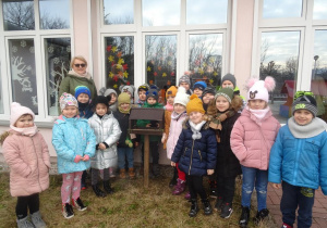 Grupa dzieci z panią Arletką stoją wokół karmnika na dworze.